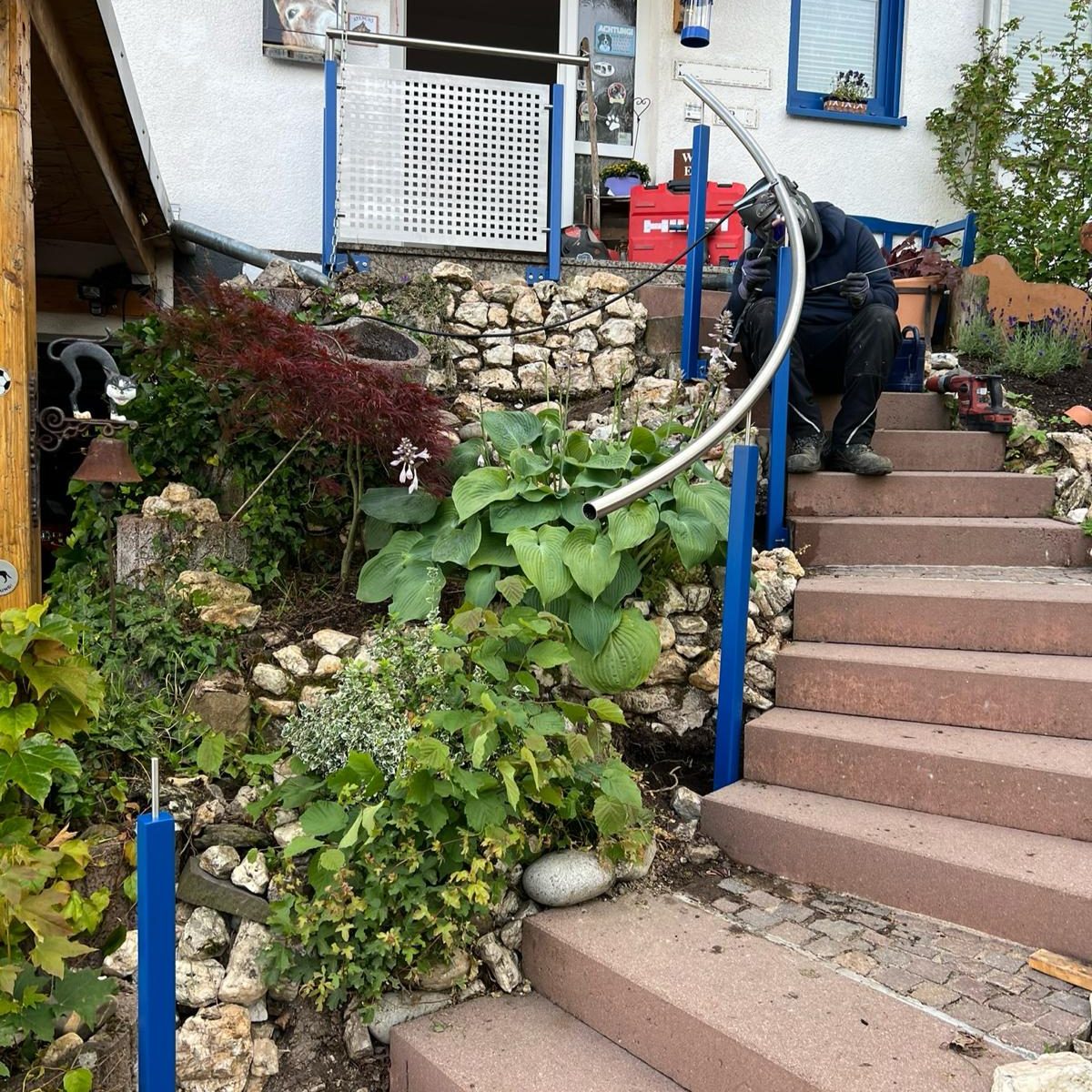 Handlauf, Gebogen, Rundrohr, 1.4301/1.4307, Edelstahl, Baustahl, Blau, RAL-Farbe, Montage Stufen mit Pflanze und Handlauf vor einem Haus, Person sitzt auf den Stufen.
